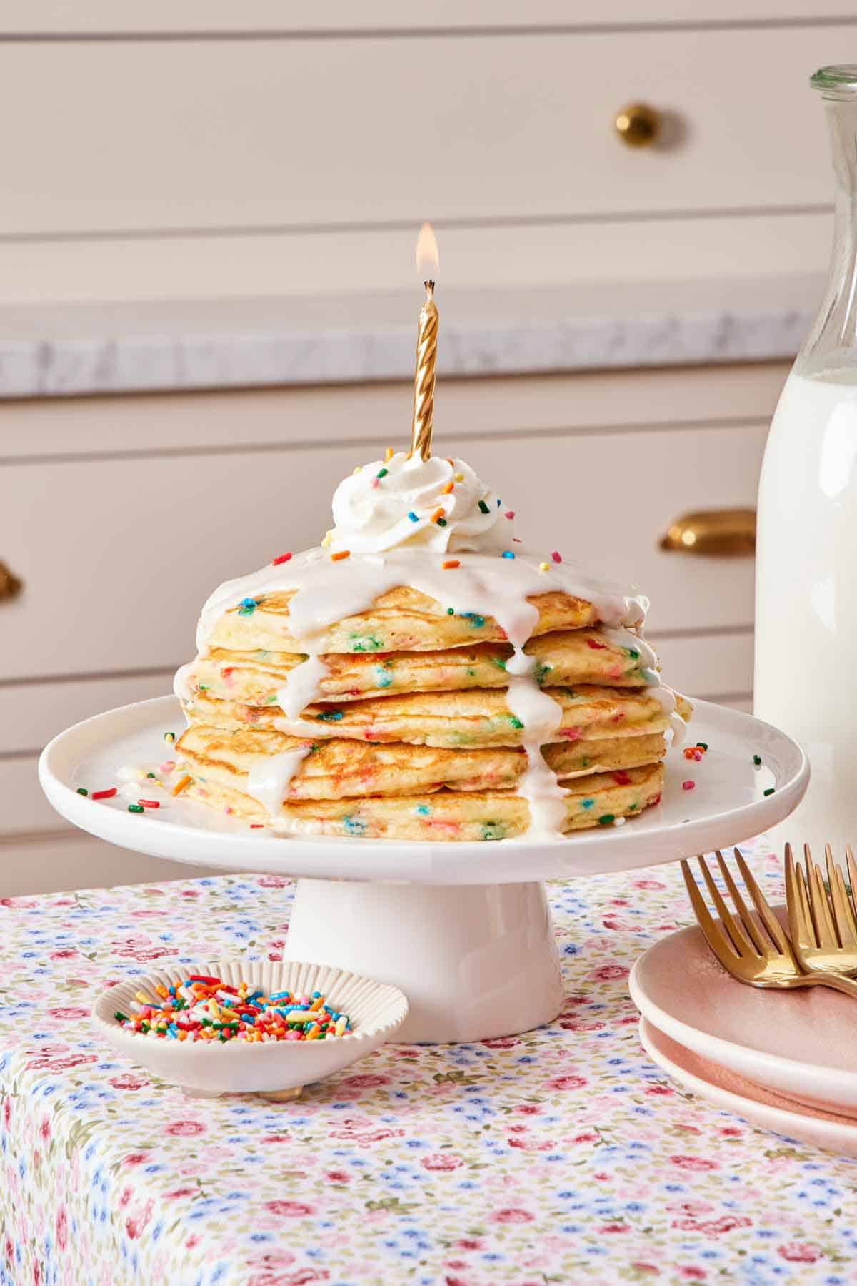 A small cake stand with a stack of funfetti pancakes with icing, whipped cream, and a lit candle on top. A bowl of sprinkles, stack of plates, forks, and milk off to the side.