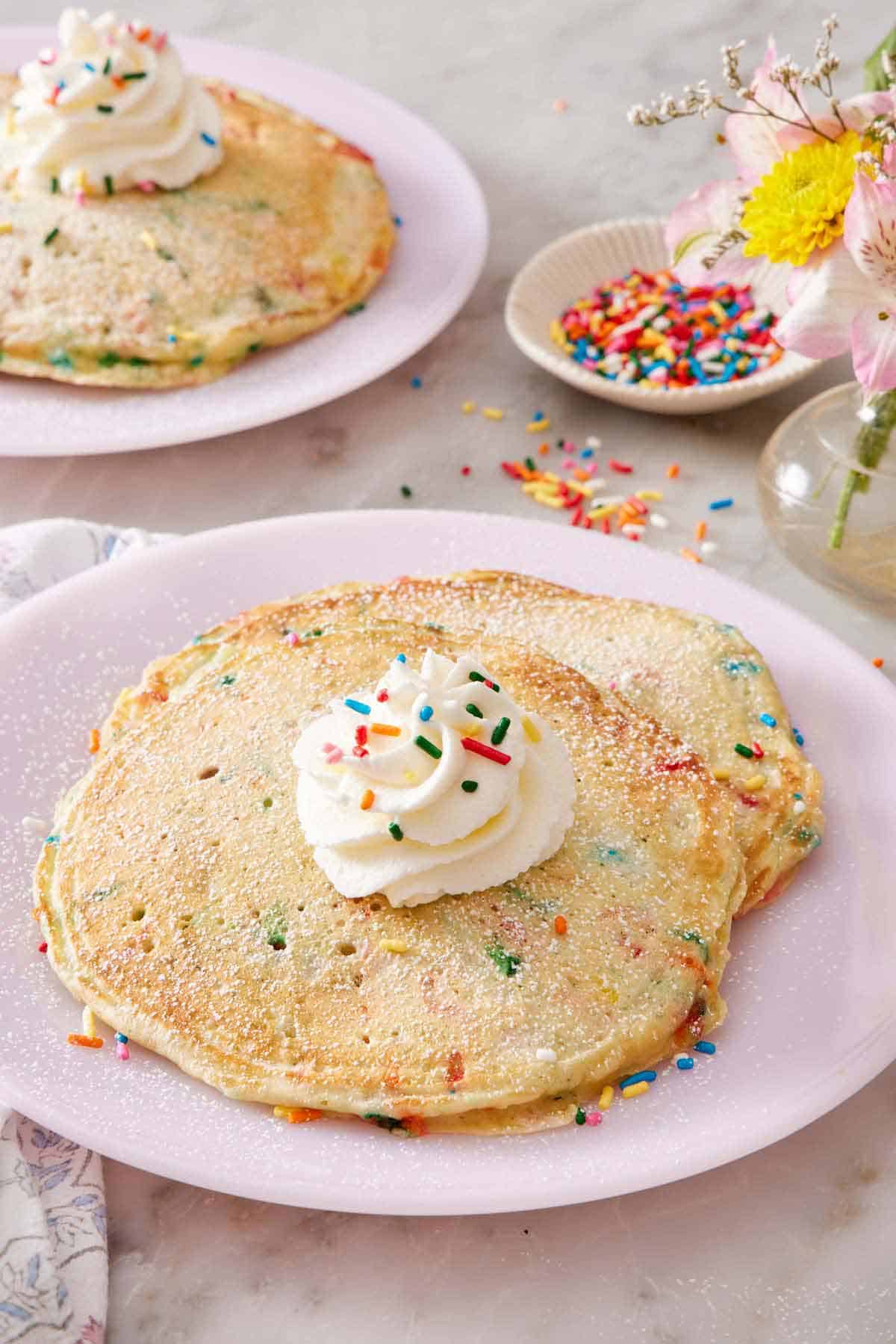 A plate of two funfetti pancakes with a dollop of whipped cream on top with sprinkles. More pancakes in the background along with a bowl of sprinkles and vase of flowers in.