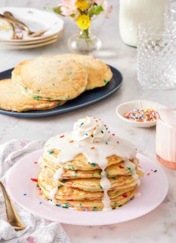 A stack of funfetti pancakes with icing, whipped cream, and sprinkles on top. More pancakes in the background along with a bowl of sprinkles, glasses, plates, forks, and flowers.