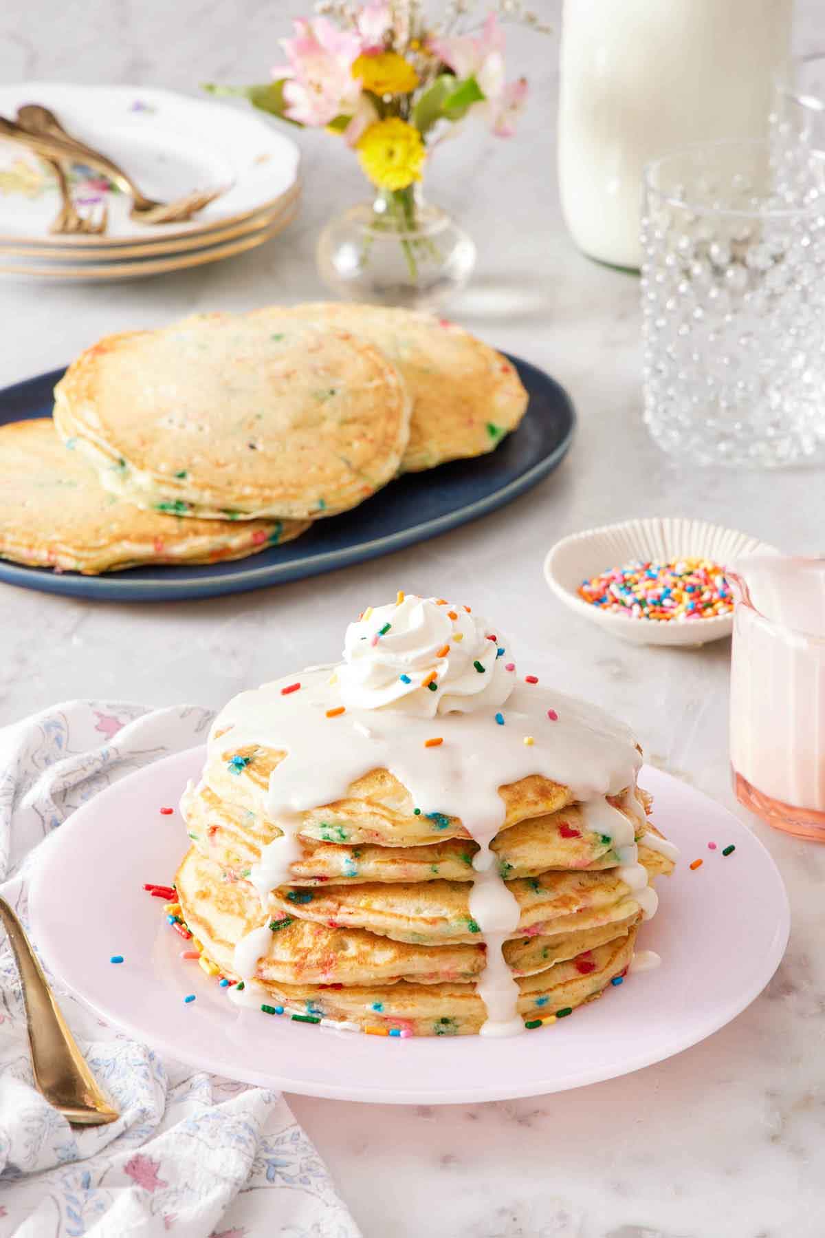 A stack of funfetti pancakes with icing, whipped cream, and sprinkles on top. More pancakes in the background along with a bowl of sprinkles, glasses, plates, forks, and flowers.