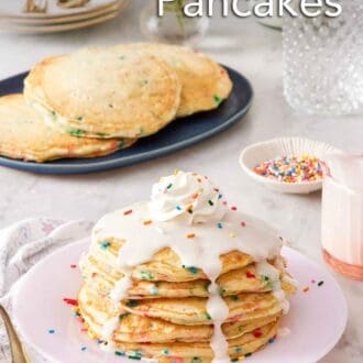 Pinterest graphic of a stack of funfetti pancakes with icing, whipped cream, and sprinkles on top. More pancakes in the background along with a bowl of sprinkles, glasses, plates, forks, and flowers.