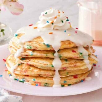 A stack of funfetti pancakes with icing, whipped cream, and sprinkles on top. More sprinkles on the side with a fork and flowers.