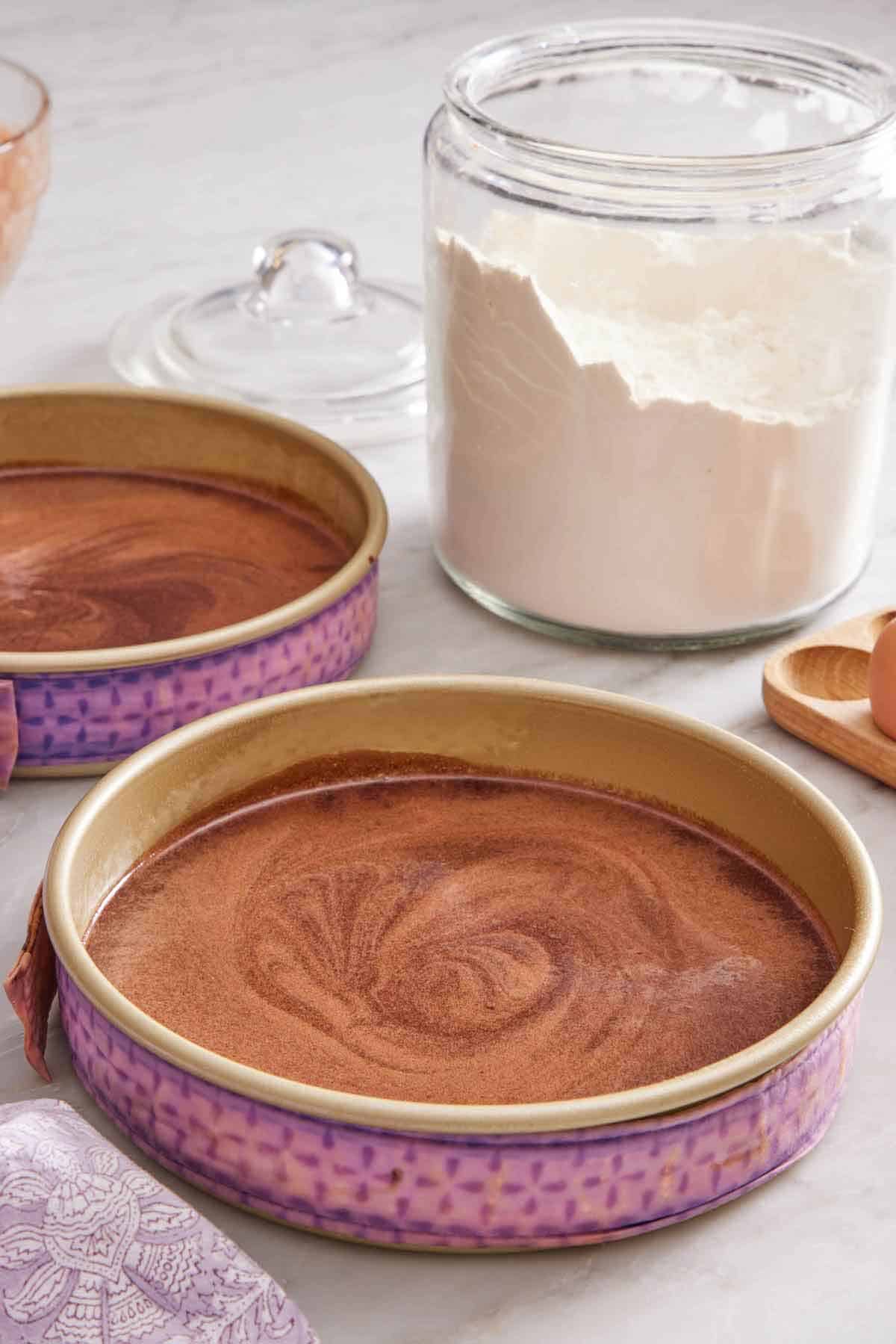 Two baking pans with chocolate cake batter with fabric cake strips wrapped around them and a jar of flour in the background.