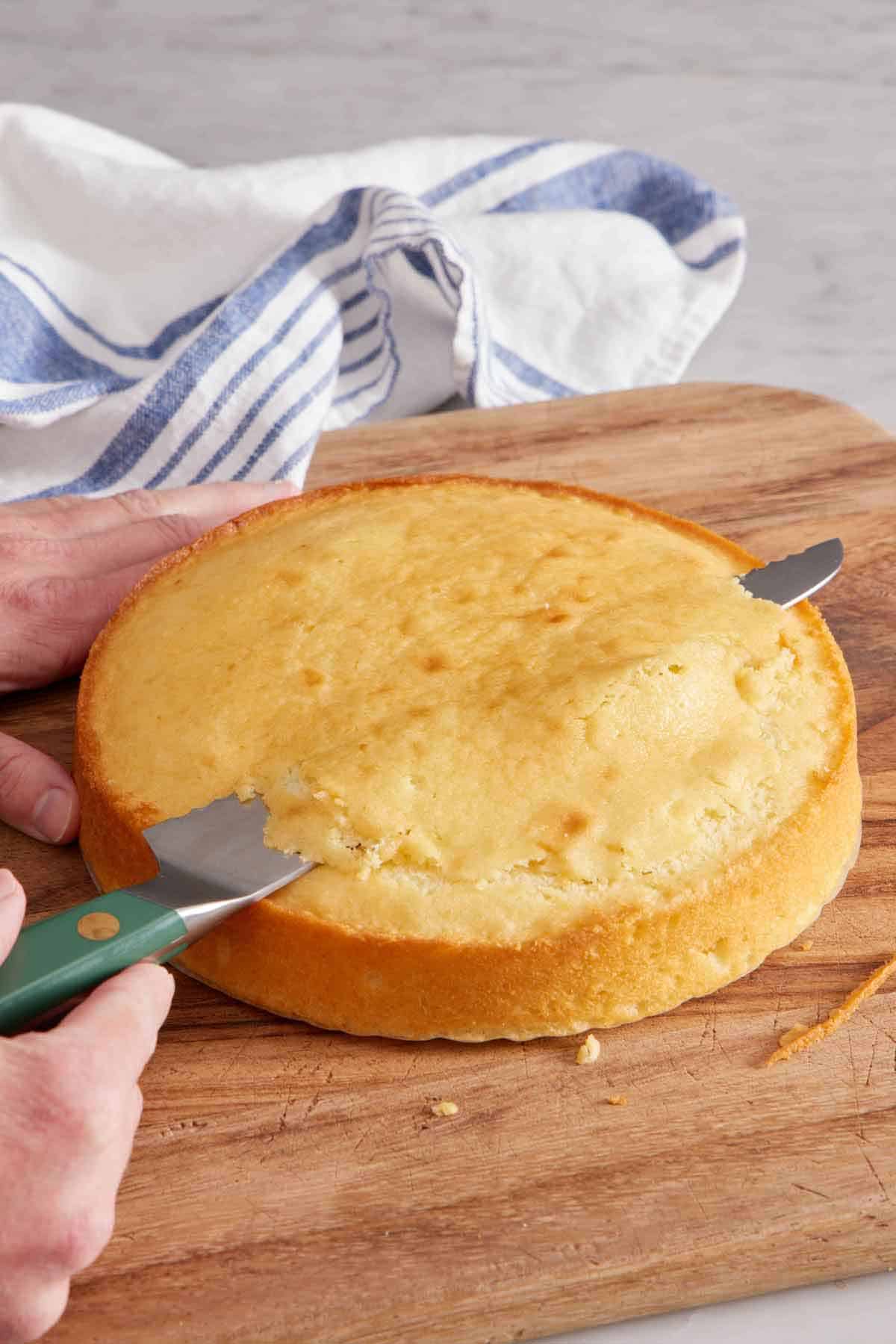 A serrated knife cutting the dome off the top of a cake on a wooden cutting board.