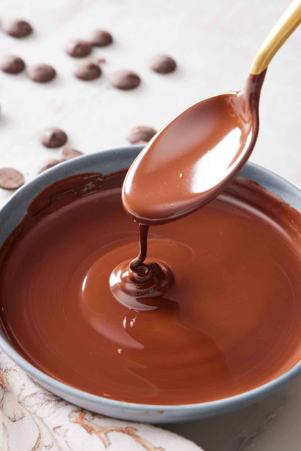 A spoon lifted from a bowl of melted chocolate with some drizzling off the spoon. Chocolate chips scattered in the background.