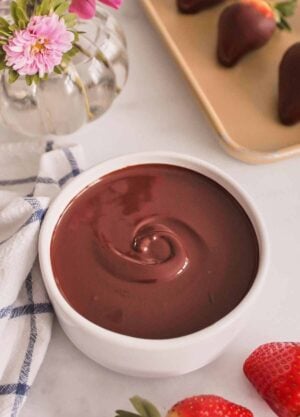 A bowl of melted chocolate. Strawberries scattered on the side and a vase of flowers in the back along with chocolate dipped strawberries on a sheet pan.