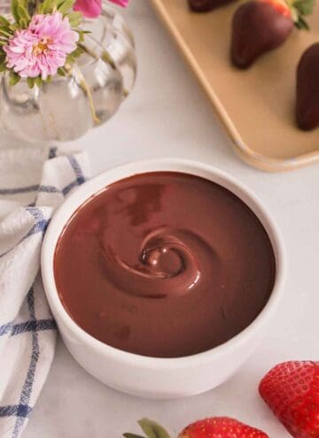 A bowl of melted chocolate. Strawberries scattered on the side and a vase of flowers in the back along with chocolate dipped strawberries on a sheet pan.