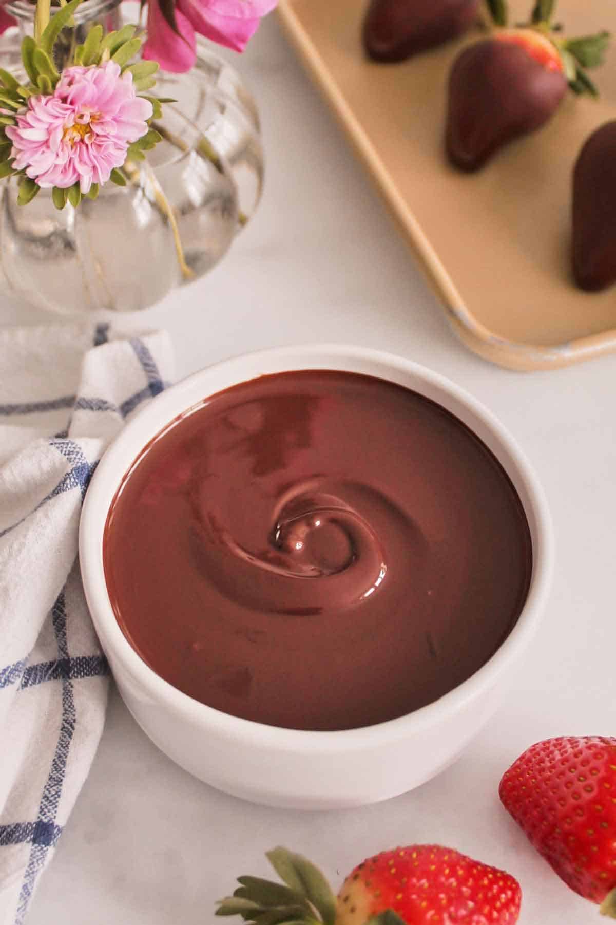 A bowl of melted chocolate. Strawberries scattered on the side and a vase of flowers in the back along with chocolate dipped strawberries on a sheet pan.