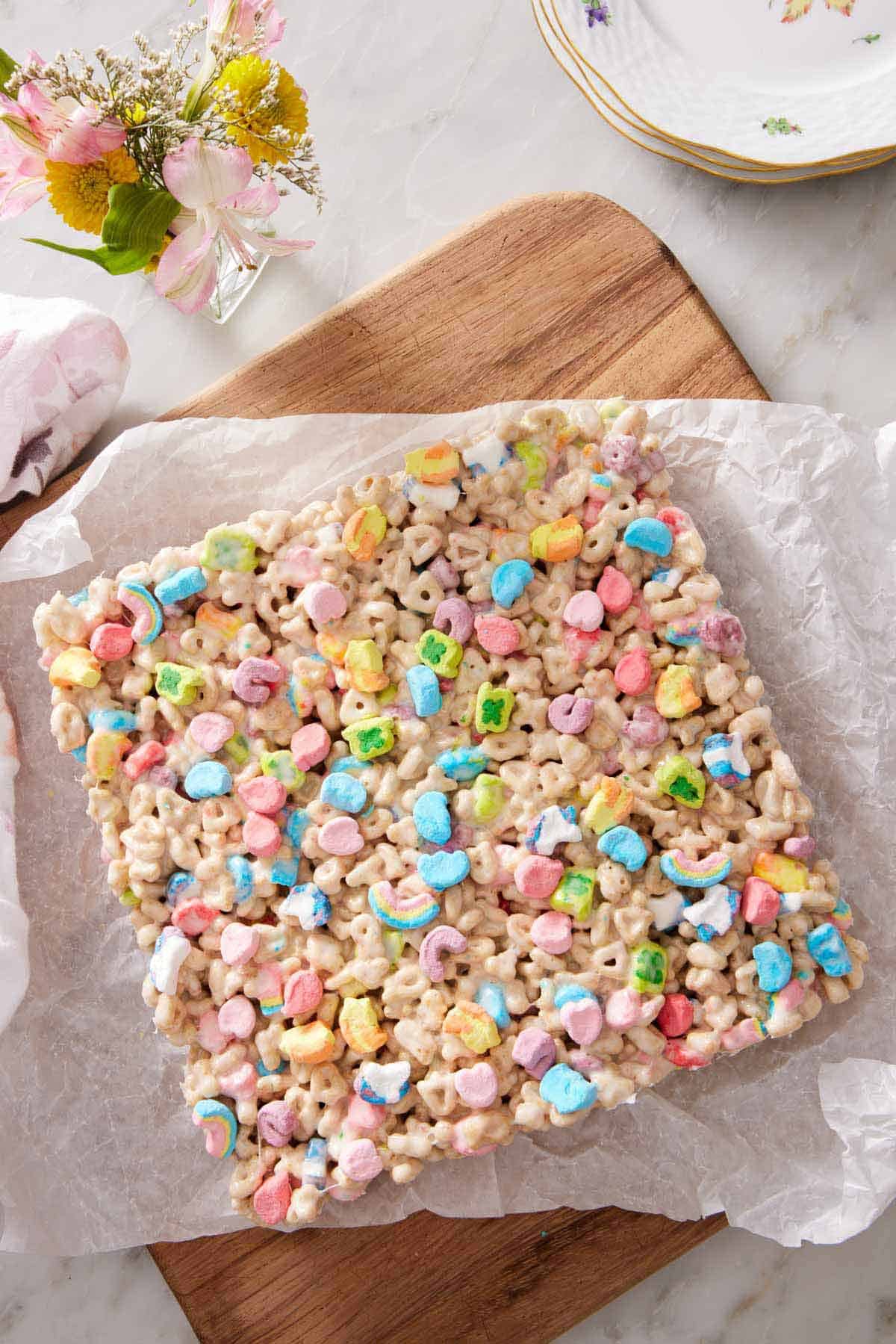 Overhead view of a slab of uncut Lucky Charms treats on a parchment-lined serving board. Flowers and plates on the side.