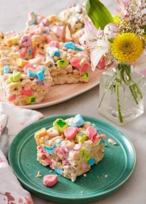 A square piece of Lucky Charms treat on a plate. A platter with more Lucky Charms treats in the background along with a vase of flowers.