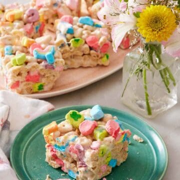A square piece of Lucky Charms treat on a plate. A platter with more Lucky Charms treats in the background along with a vase of flowers.