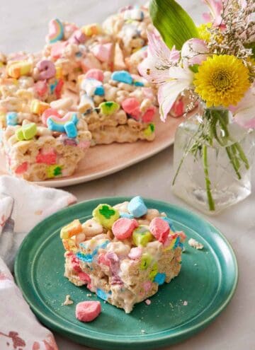 A square piece of Lucky Charms treat on a plate. A platter with more Lucky Charms treats in the background along with a vase of flowers.