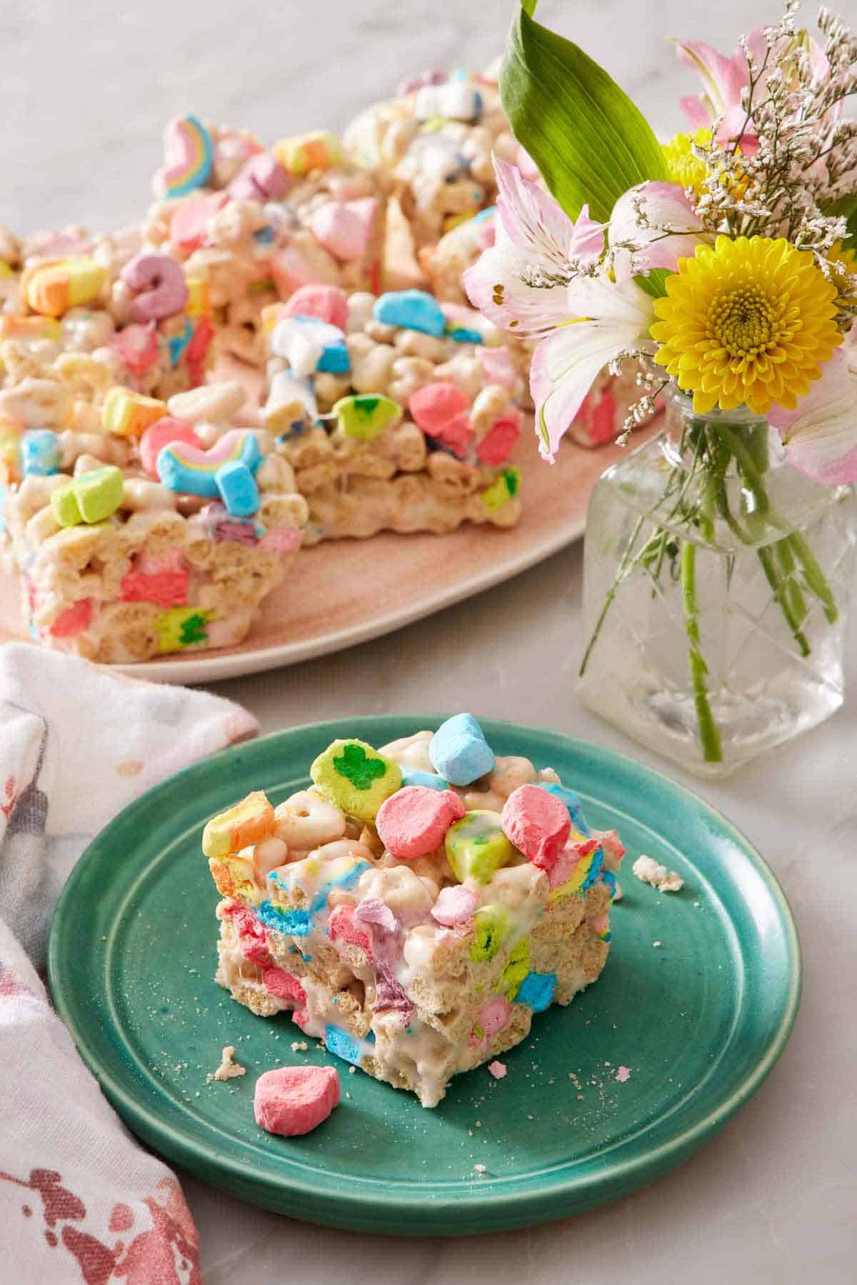 A square piece of Lucky Charms treat on a plate. A platter with more Lucky Charms treats in the background along with a vase of flowers.