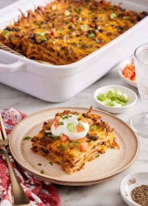 A plate with a serving of Mexican lasagna with a baking dish in the background with the rest of the lasagna. Bowls of garnishes and a drink on the side.