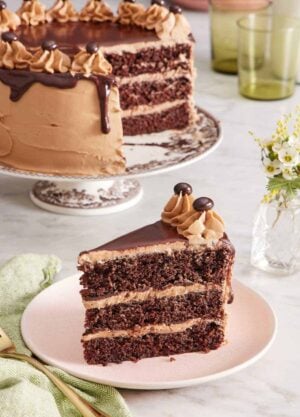 A slice of moist mocha cake on a plate with the rest of the cake on a cake stand in the background. Vase of flowers on the side.