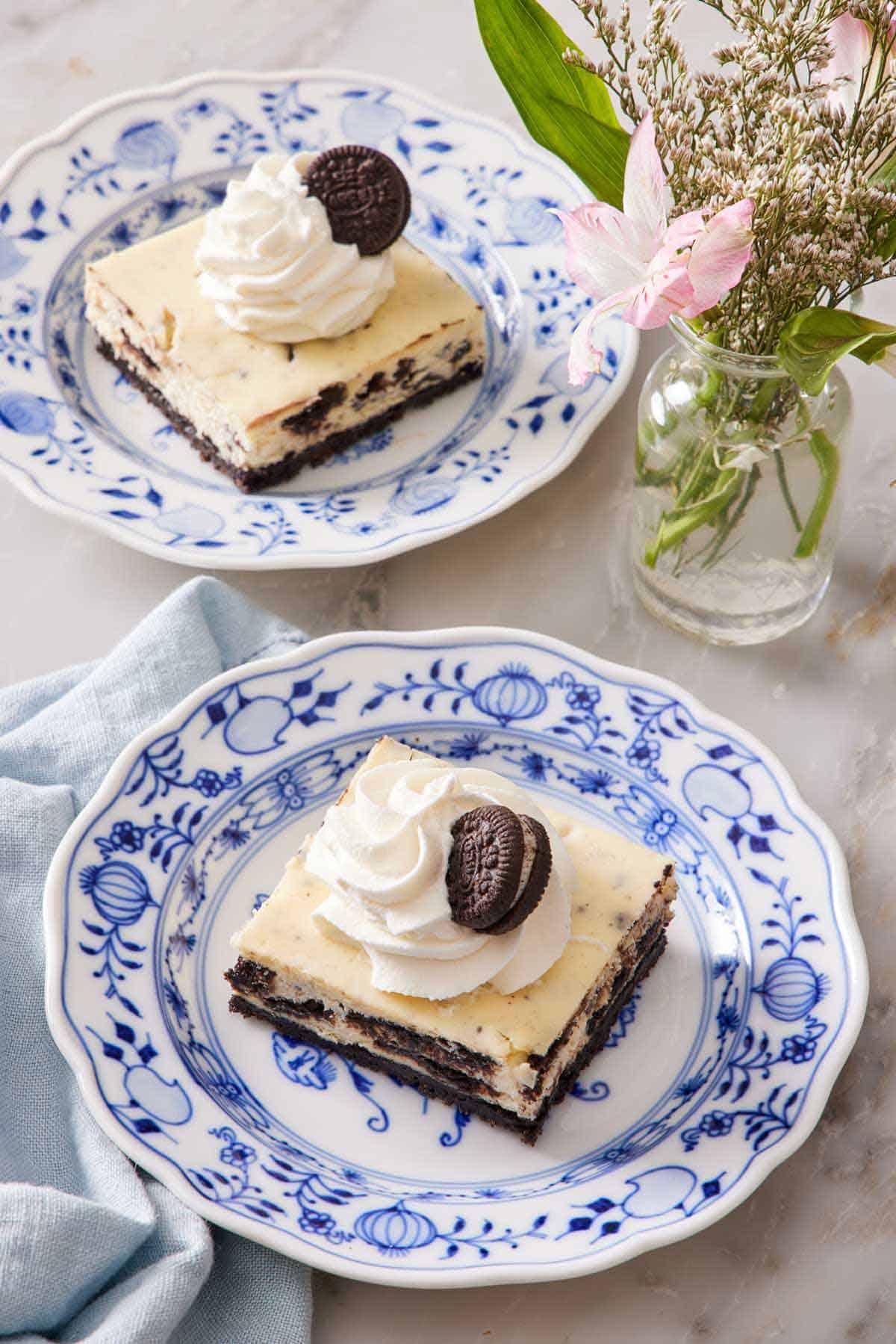 Two plates with Oreo cheesecake bars topped with whipped cream and an Oreo cookie. Flowers in a small vase between the plates.