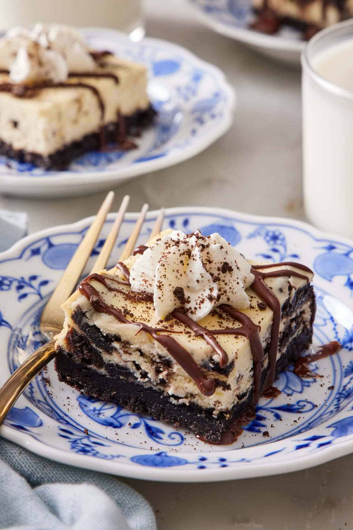 A piece of Oreo cheesecake bar on a plate with chocolate drizzled on top and a dollop of whipped cream. A fork on the plate. Another plated serving in the back with a glass of milk.