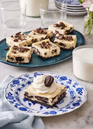 A square piece of Oreo cheesecake bar on a plate with a platter with more in the background along with a glass of milk.