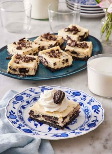 A square piece of Oreo cheesecake bar on a plate with a platter with more in the background along with a glass of milk.