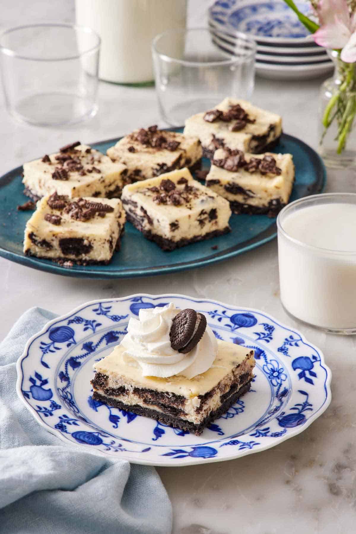 A square piece of Oreo cheesecake bar on a plate with a platter with more in the background along with a glass of milk.