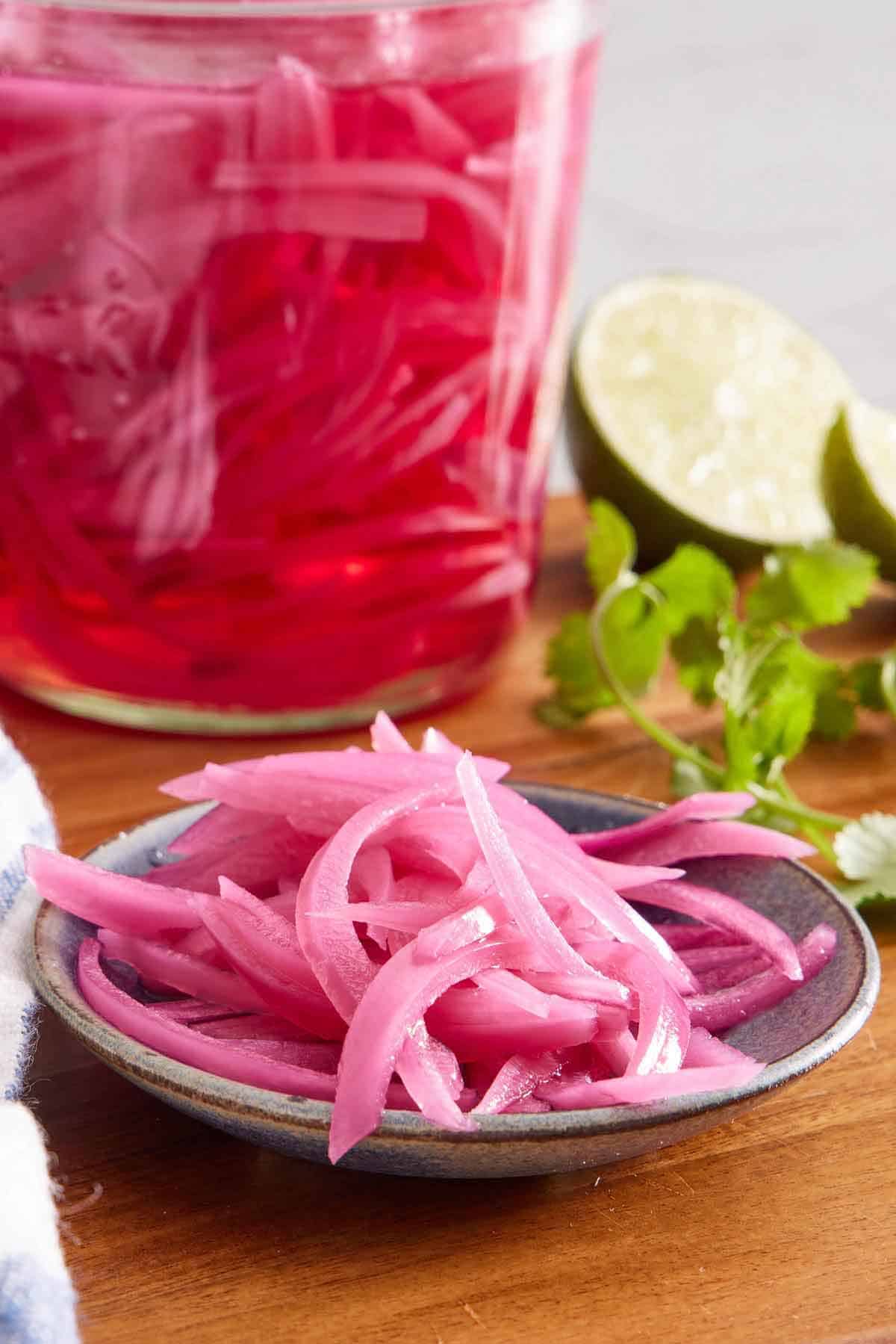 A plate of pickled onions on a wooden serving board with a jar of more pickled onions in the background along with cut limes and cilantro.