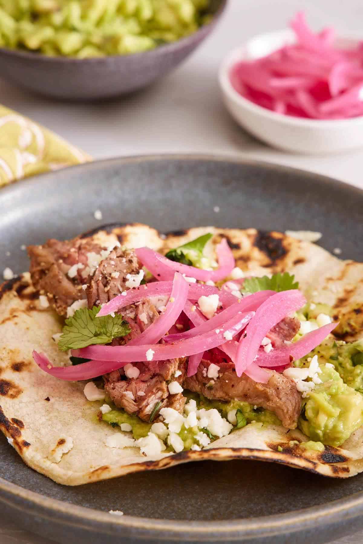 Pickled onions on top of a steak taco on a plate. A bowl of more pickled onions in the background along with guacamole.