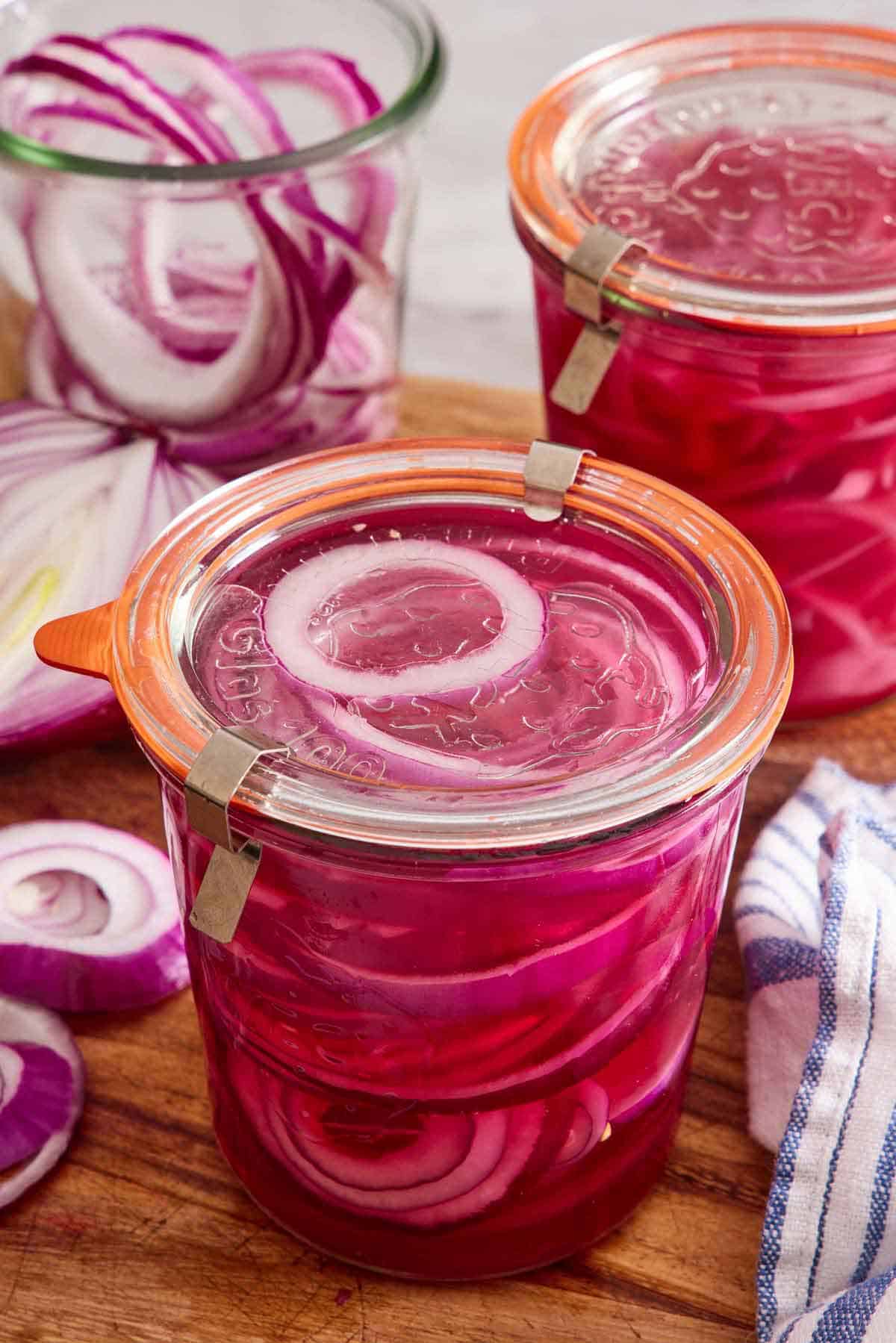 Two weck jars of pickled onions with their lids on. Another jar in the back with red onions placed inside.