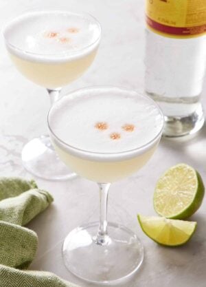 Two coupe glasses of pisco sour with cut limes and a bottle of pisco in the background.
