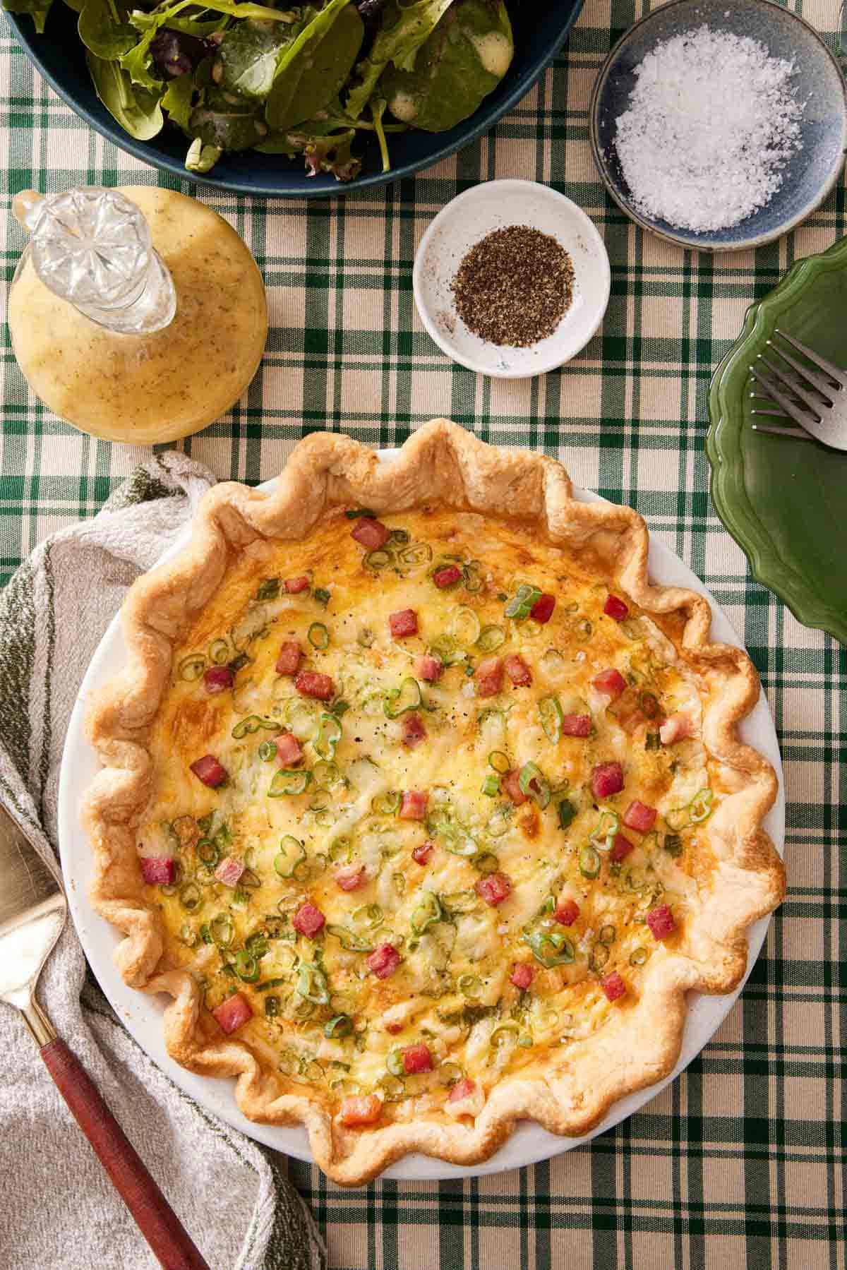 Overhead view of a quiche with ham and green onions. A bowl of salt, pepper, salad dressing, and salad on the side.