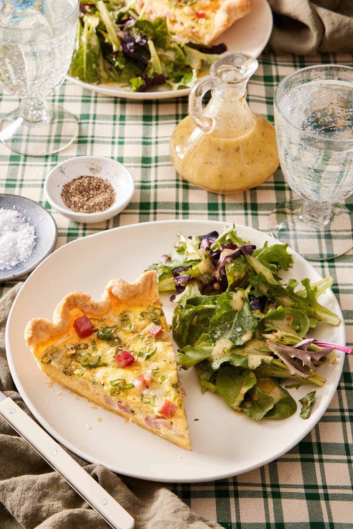 A plate with a piece of quiche with some salad topped with dressing. Salt, pepper, salad dressing, and drinks in the background.