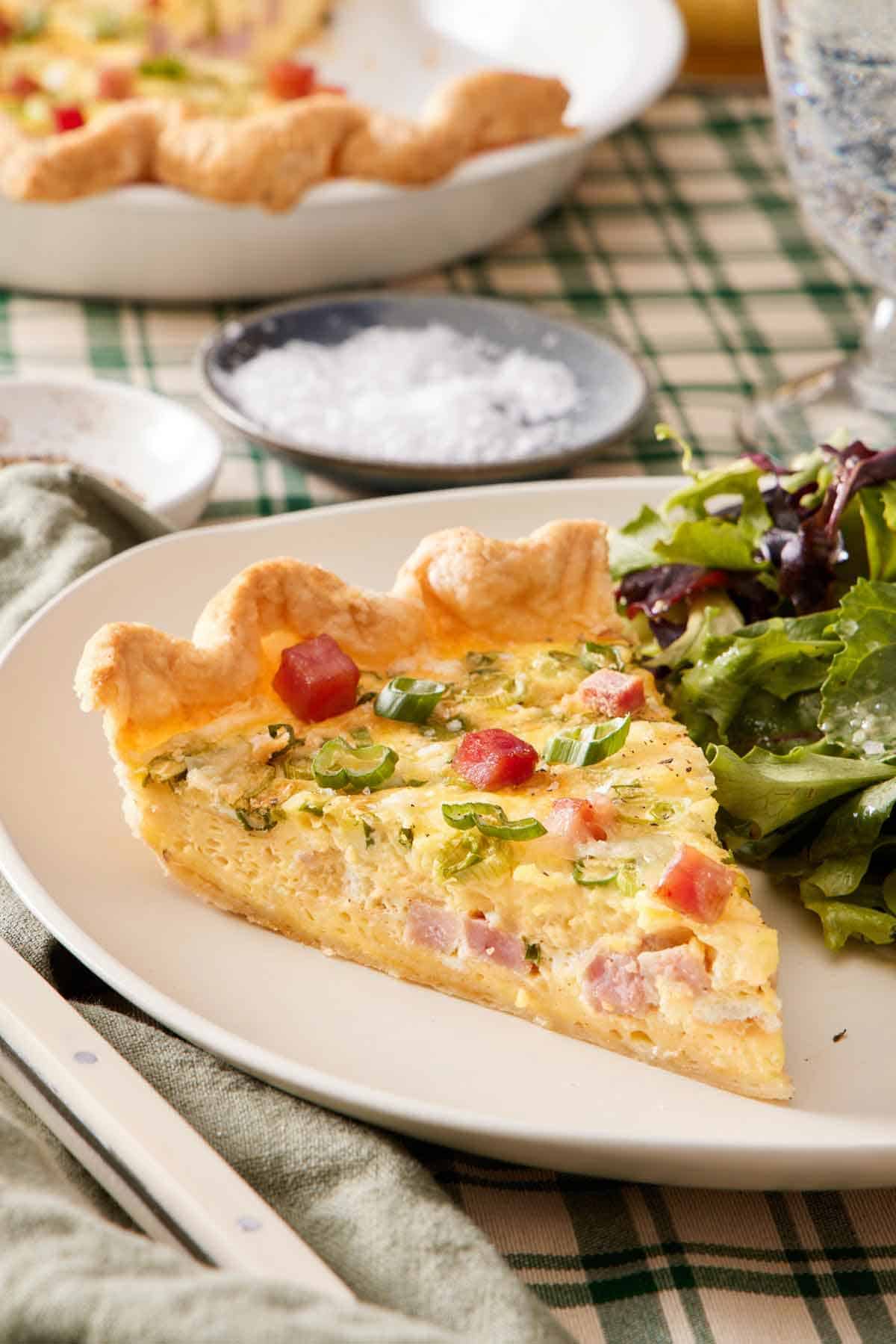 A close up view of a slice of quiche with salad on the side. A bowl of salt in the background with more quiche.