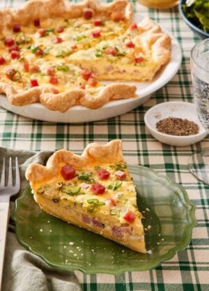 A slice of quiche on a green plate with the rest of the quiche in the background along with a bowl of pepper.