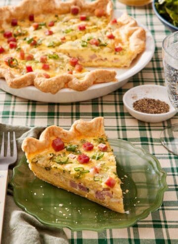 A slice of quiche on a green plate with the rest of the quiche in the background along with a bowl of pepper.