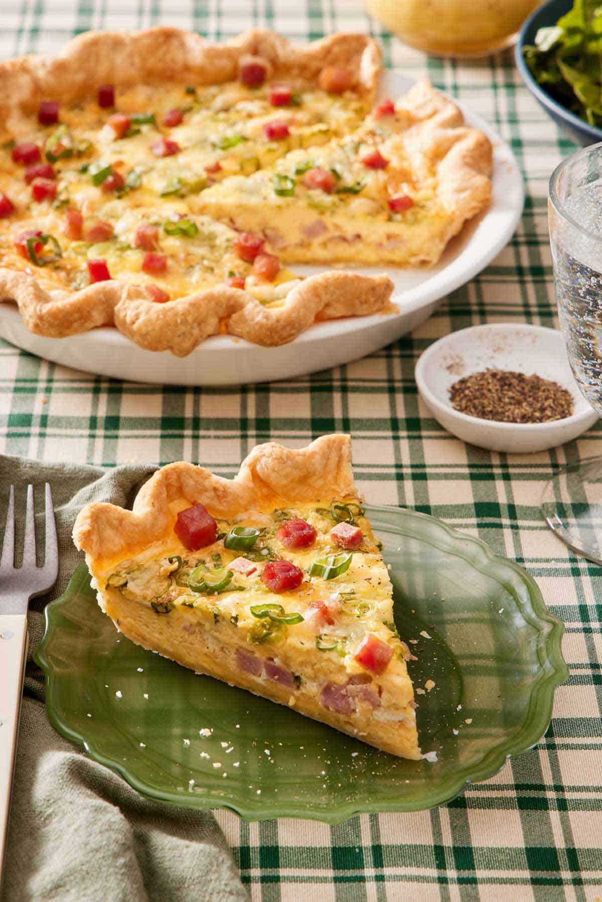 A slice of quiche on a green plate with the rest of the quiche in the background along with a bowl of pepper.