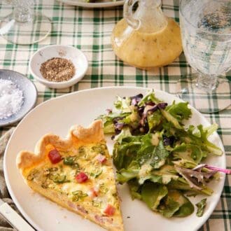 Pinterest graphic of a plate with a piece of quiche with some salad topped with dressing. Salt, pepper, salad dressing, and drinks in the back.