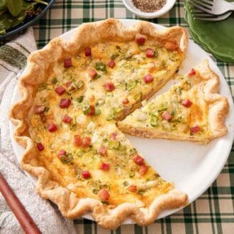 An overhead view of a quiche in the baking dish with one piece cut out and one piece removed. Salt, plates, pepper, and salad on the side.