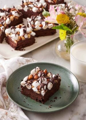 A plate with a rocky road bar with more on a platter in the background with a vase of flowers and glass of milk.