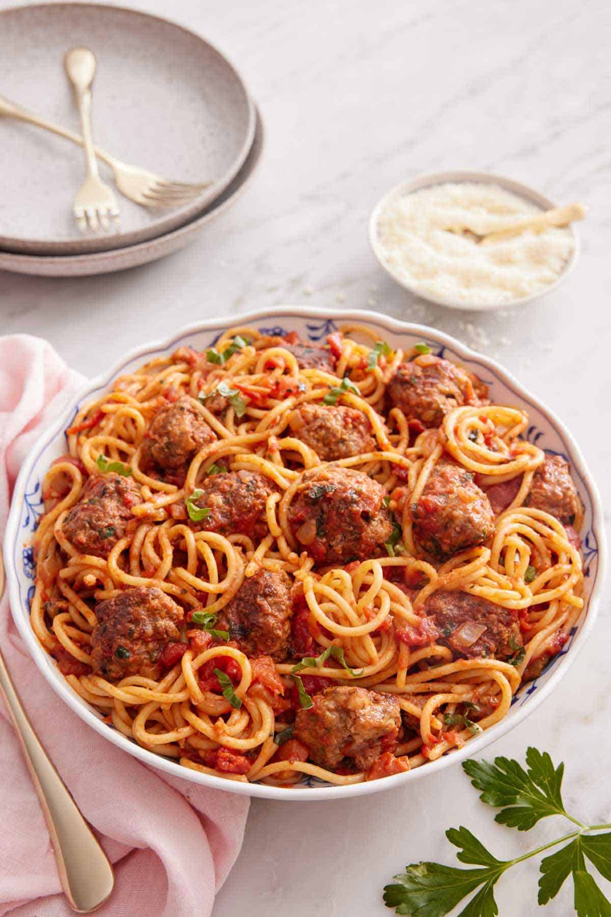 A bowl of spaghetti and meatballs topped with chopped parsley. A bowl of parmesan in the back and a stack of plates with forks.