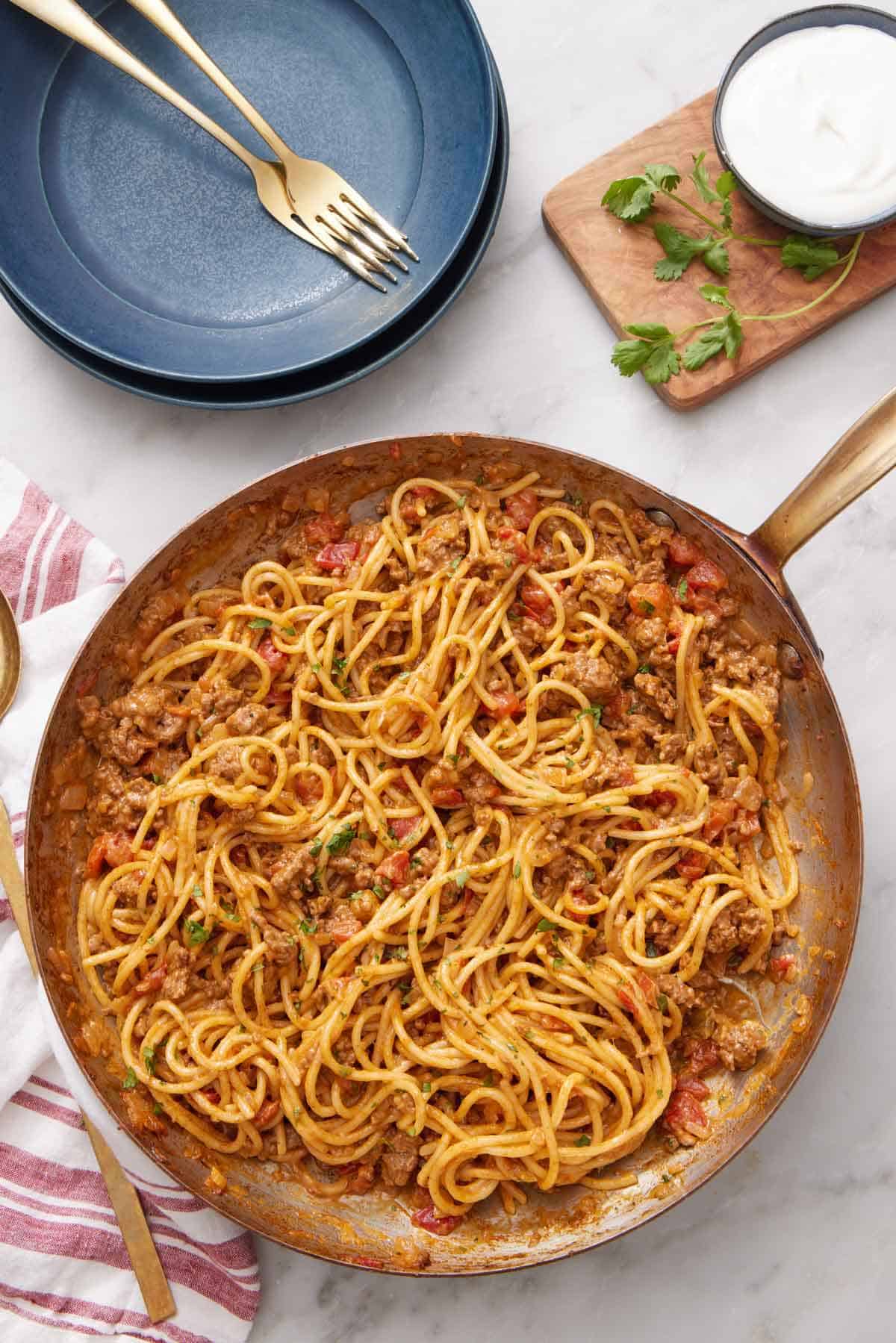 Overhead view of a skillet of taco spaghetti. A stack of plates, forks, cilantro, and sour cream on the side.