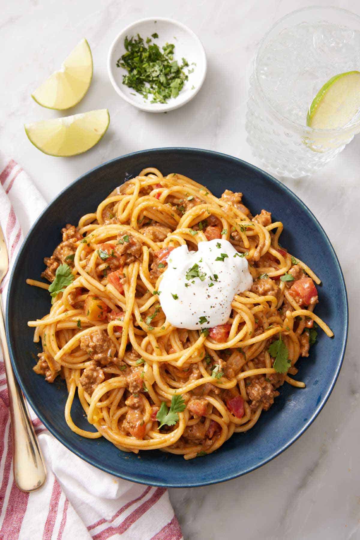 Overhead view of a bowl of taco spaghetti with a dollop of sour cream and chopped cilantro. Cilantro, lime wedges, and a drink beside it.