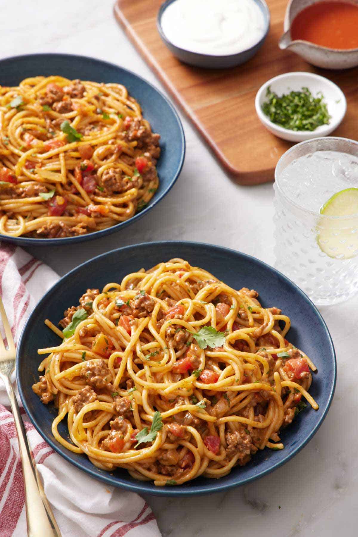 Two bowls of taco spaghetti with a drink, bowl of chopped cilantro, and two sauces in the background.