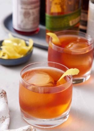 Two glasses of Vieux Carré cocktails with lemon peel garnish. A bowl of more lemon peels in the back along with bottles of alcohol.
