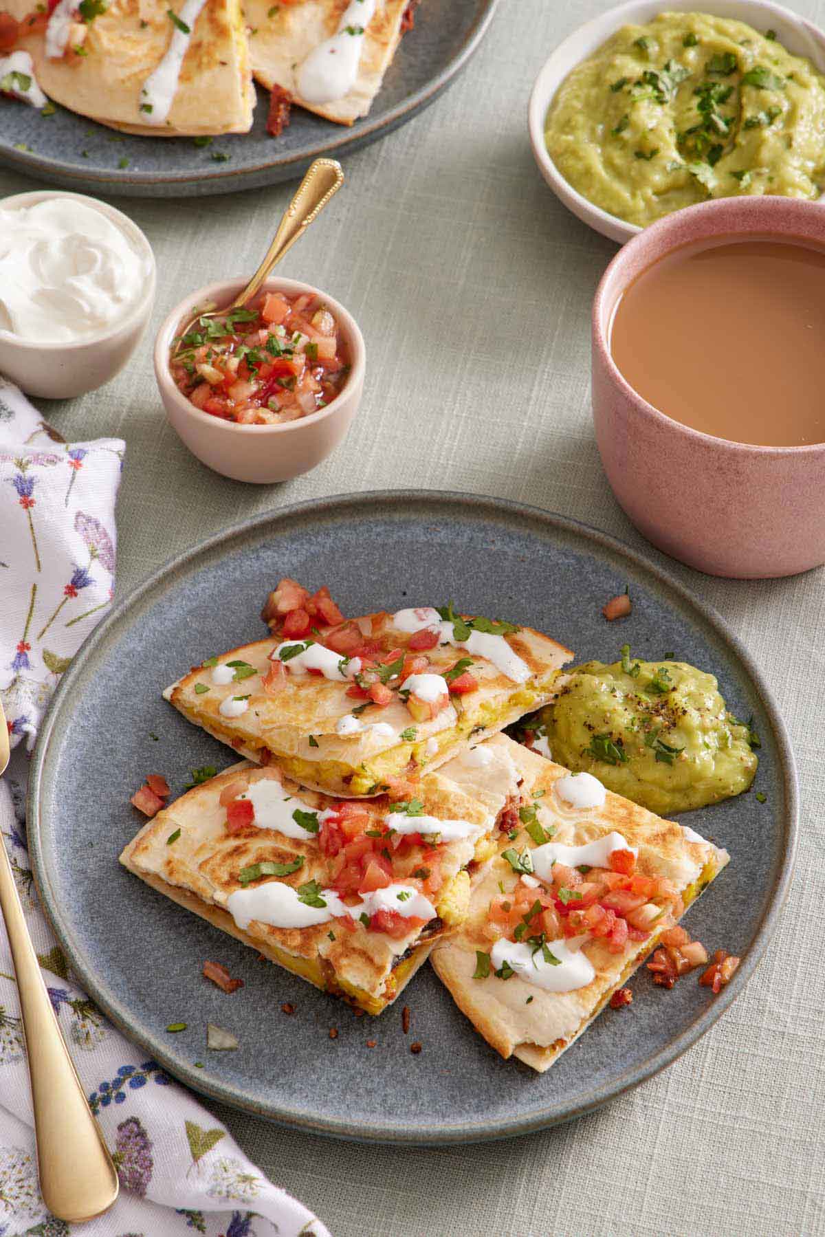 A plate with three pieces of breakfast quesadillas topped with pico de gallo, sour cream, and guacamole. More toppings in bowls in the background along with a cup of coffee.
