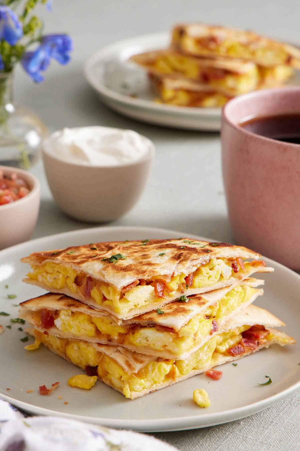 A slightly profile view of a stack of three breakfast quesadillas. Bowls of sour cream and pico de gallo and coffee in the back.