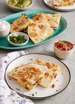 A plate with two pieces of breakfast quesadillas. A platter in the background with more with bowls of sour cream and chopped cilantro. A bowl of pico de gallo and guacamole in the background.