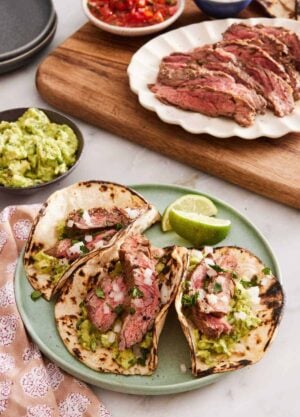 Three carne asada tacos on a plate with lime wedges. Guacamole, sliced grilled meat, pico de gallo, onions on a wooden serving board.