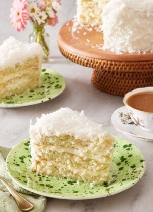 A slice of coconut cake on a plate. In the background is another plated slice and the rest of the cake on a cake stand.