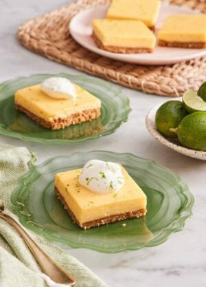 Two plates with key lime pie bars topped with whipped cream and lime zest. More slices in the background along with a plate of key limes.