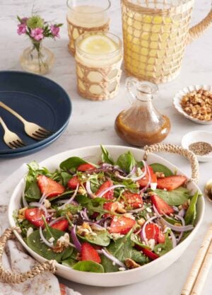 A large platter of strawberry salad. A pitcher of lemonade in the background with glasses, salad dressing, flowers, plates, and forks.