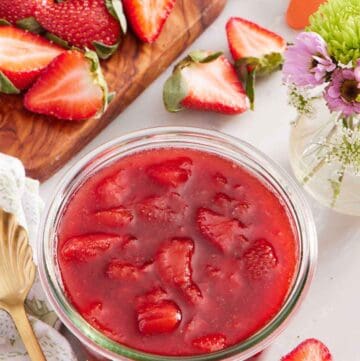 A jar of strawberry sauce. Cut strawberries scattered around and on a cutting board along with a vase of flowers.